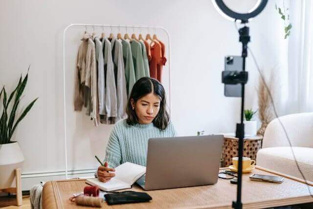 A woman creating online content on her laptop with a clothes rack behind her.
