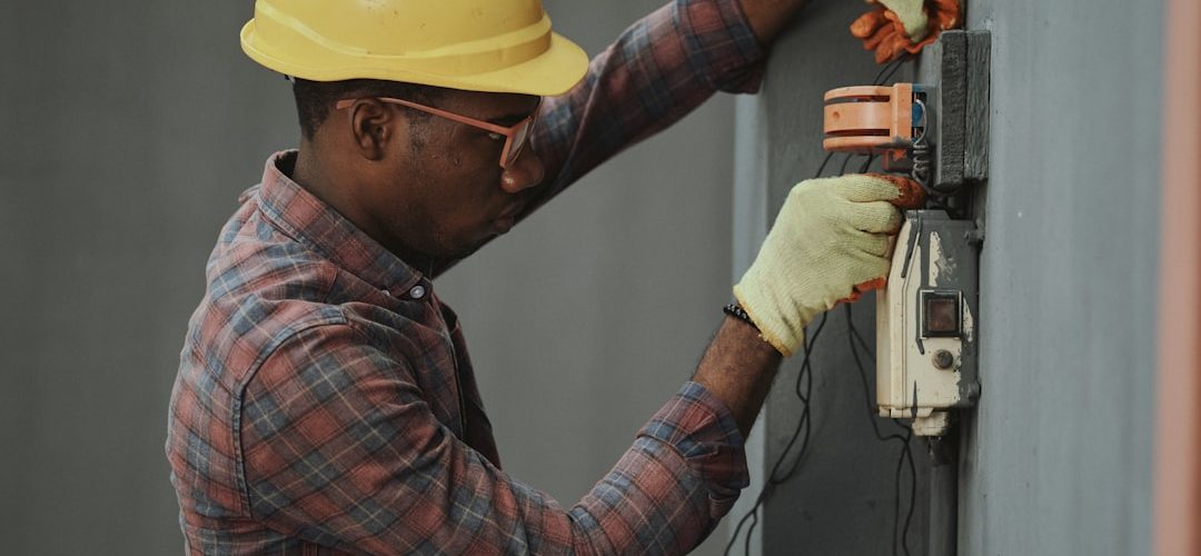 man in brown and white plaid dress shirt and yellow hard hat holding black and orange technician installation wall mounted intercom safety equipment