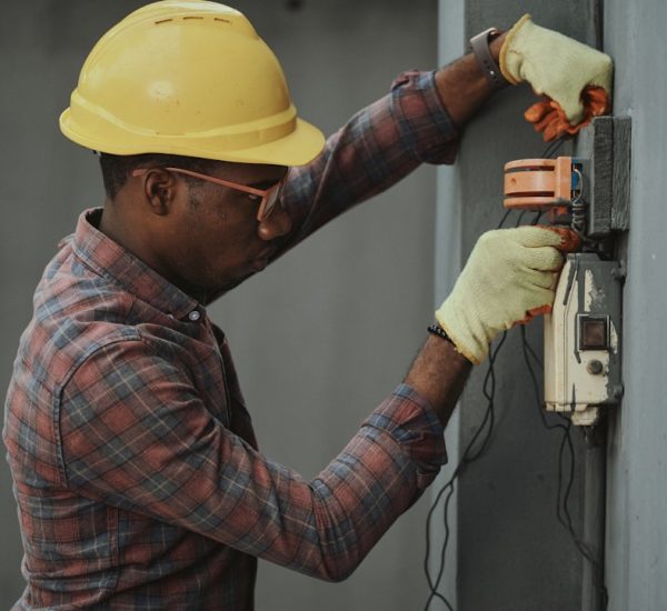 man in brown and white plaid dress shirt and yellow hard hat holding black and orange technician installation wall mounted intercom safety equipment