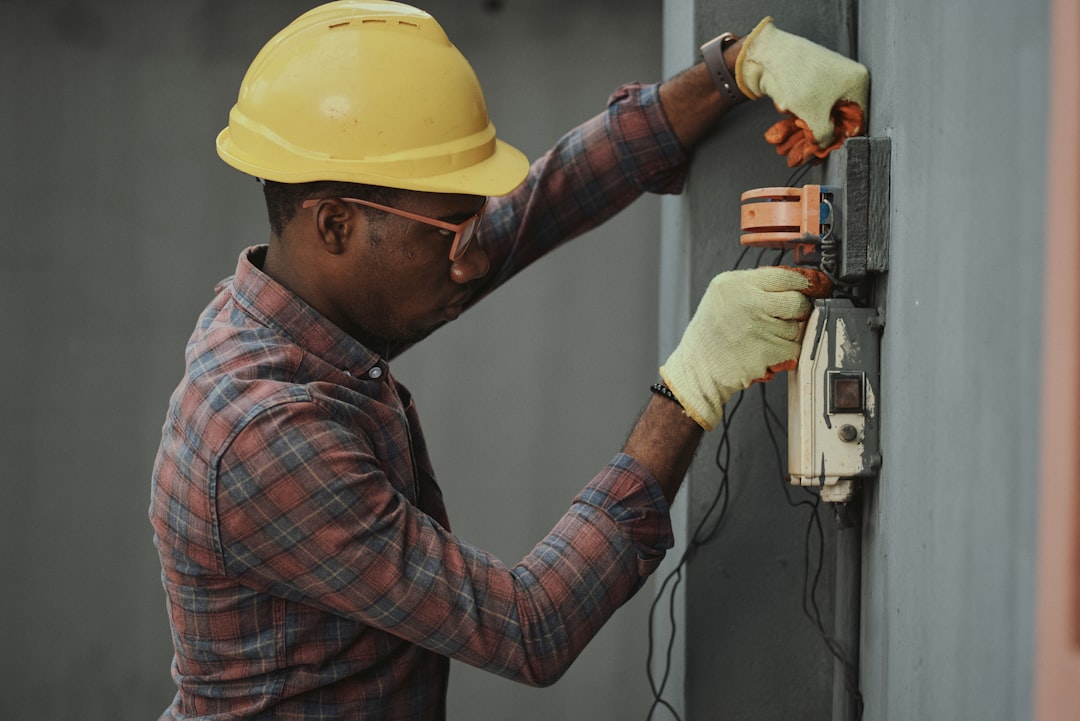 man in brown and white plaid dress shirt and yellow hard hat holding black and orange technician installation wall mounted intercom safety equipment