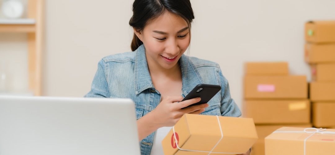 woman checking package with phone near laptop and boxes online store analytics mobile commerce