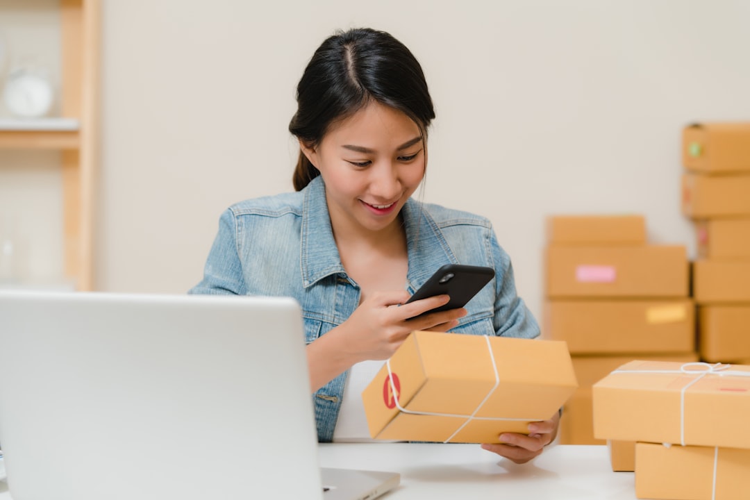 woman checking package with phone near laptop and boxes online store analytics mobile commerce